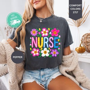 May include: A charcoal gray short-sleeve t-shirt featuring the word NURSE in colorful, patterned block letters surrounded by a vibrant illustration of various pink, purple, white, and yellow flowers with green stems and leaves.