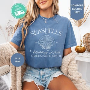 May include: A blue jean colored t-shirt featuring a white graphic of a seashell centered between the text SEASHELLS AND WEDDING BELLS CLAIRE'S BACHELORETTE. The shirt has a crew neckline and short sleeves, with the text COMFORT COLORS 1717 visible.