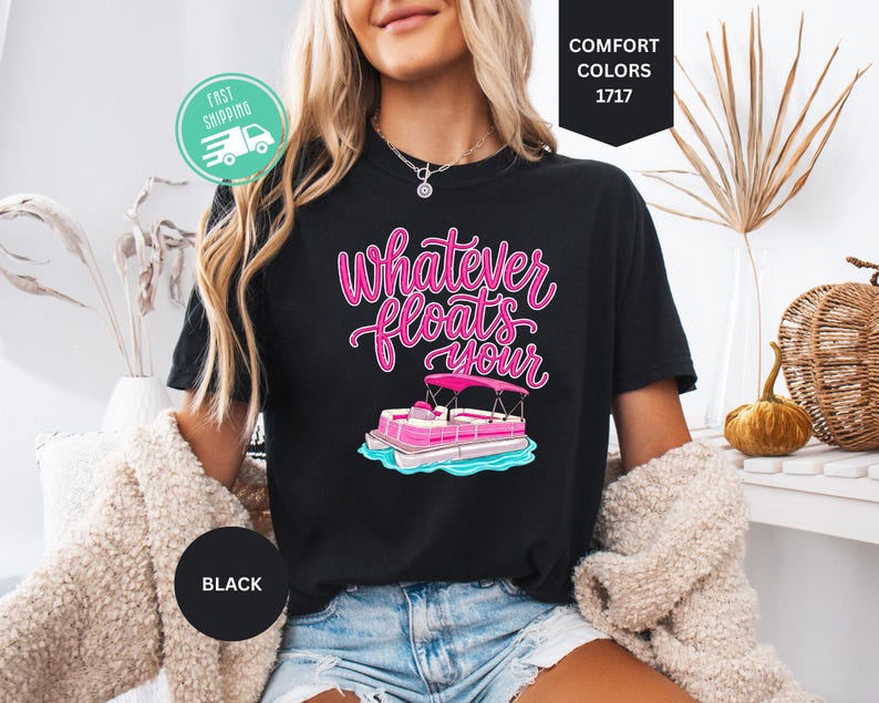 Op de afbeelding: Zwart T-shirt met de roze tekst "Whatever Floats Your" en een roze en witte pontonbootafbeelding. Het shirt is in Comfort Colors 1717-stijl.
