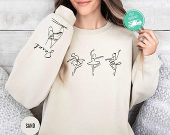 Könnte beinhalten: Ein sandfarbener Pullover mit einer Grafik von drei Tänzerinnen in einer Reihe. Der Text "Dance" steht auf dem linken Ärmel.