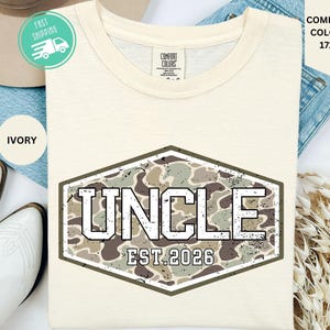 May include: An ivory-colored t-shirt featuring a hexagonal graphic with a camouflage pattern. The text "UNCLE" is printed in large, bold, white block letters, with "EST. 2026" in smaller white text underneath, all centered on the chest.