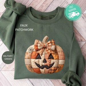 Op de afbeelding: Een militair groen sweatshirt met een centrale faux patchwork pompoenafbeelding. De pompoen heeft een jack-o'-lantern gezicht, een geruite strik aan de bovenkant en diverse oranje en beige stofpatronen met spinnenwebontwerpen.