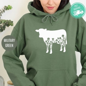 K&ouml;nnte beinhalten: Ein gr&uuml;ner Kapuzenpullover mit einer wei&szlig;en Silhouette einer Kuh, in der Blumen wachsen.