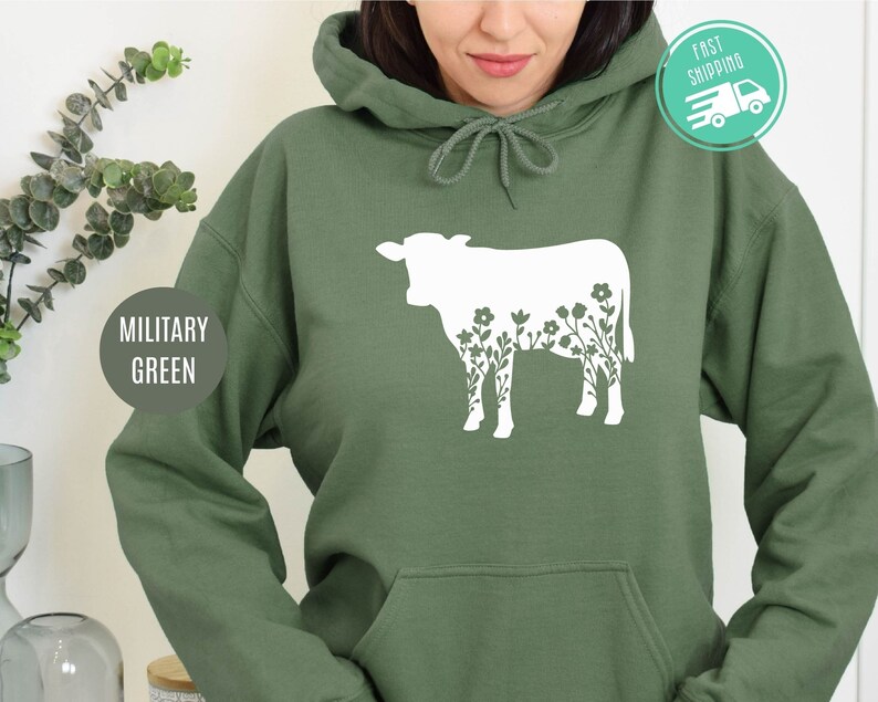 K&ouml;nnte beinhalten: Ein gr&uuml;ner Kapuzenpullover mit einer wei&szlig;en Silhouette einer Kuh, in der Blumen wachsen.
