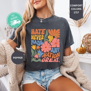 May include: A charcoal gray short-sleeve t-shirt featuring a colorful graphic print with the text HATE NEVER MADE ANY NATION GREAT. The design includes stylized orange, pink, yellow, and blue flowers with green leaves on a dark fabric background.