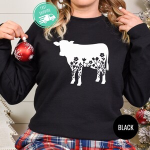 K&ouml;nnte beinhalten: Schwarzer Pullover mit einer wei&szlig;en Silhouette einer Kuh mit Blumen im Umriss. Der Text "BLACK" ist unten rechts auf dem Pullover aufgedruckt.