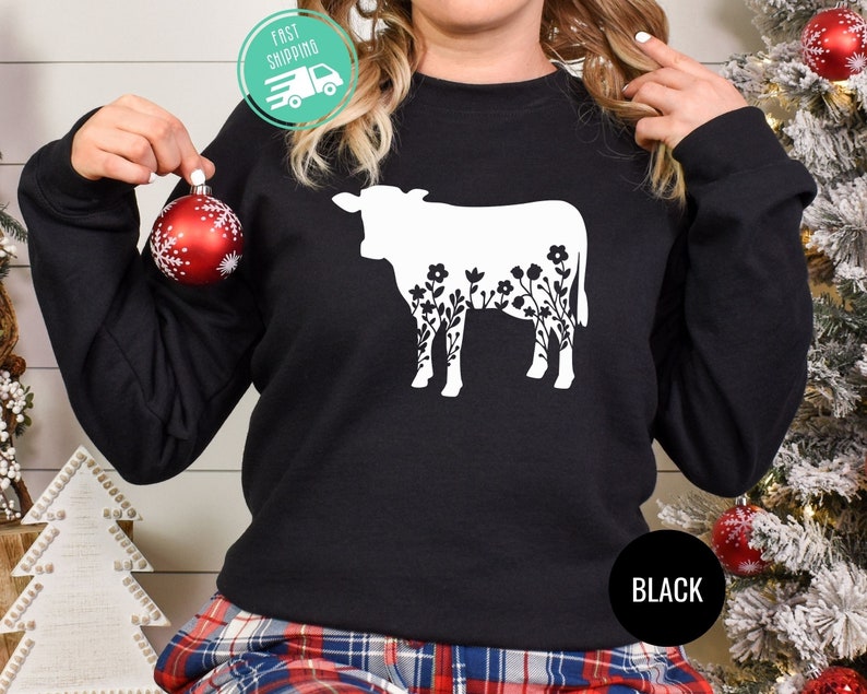 K&ouml;nnte beinhalten: Schwarzer Pullover mit einer wei&szlig;en Silhouette einer Kuh mit Blumen im Umriss. Der Text "BLACK" ist unten rechts auf dem Pullover aufgedruckt.