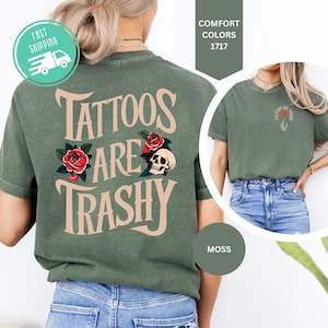 May include: A moss green t-shirt featuring the text "TATTOOS ARE TRASHY" in large beige letters on the back, accompanied by red rose and skull illustrations. A smaller version of the same graphic is printed on the front left chest area.
