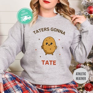 May include: A gray sweatshirt with the text "Taters Gonna Tate" and a cartoon potato with a smiling face. The sweatshirt is on a person wearing plaid pants.