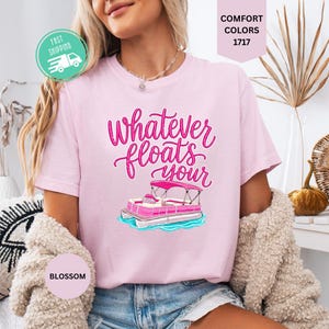 Op de afbeelding: Lichtroze T-shirt met de tekst "Whatever floats your" in roze script, met een roze pontonbootafbeelding. Het shirt heeft een ronde hals en korte mouwen. Het woord "Blossom" staat op een kleine cirkel.
