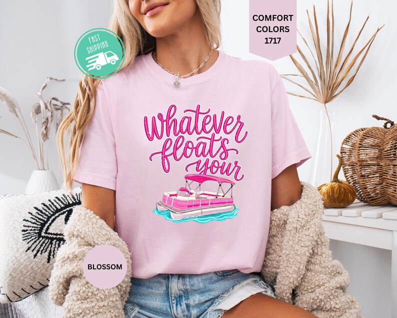 Op de afbeelding: Lichtroze T-shirt met de tekst "Whatever floats your" in roze script, met een roze pontonbootafbeelding. Het shirt heeft een ronde hals en korte mouwen. Het woord "Blossom" staat op een kleine cirkel.