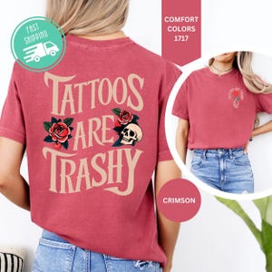 May include: A crimson short-sleeve t-shirt featuring a large graphic on the back with the text "TATTOOS ARE TRASHY" in beige, accompanied by illustrations of red roses and a skull. A smaller version of the graphic appears on the front left chest.