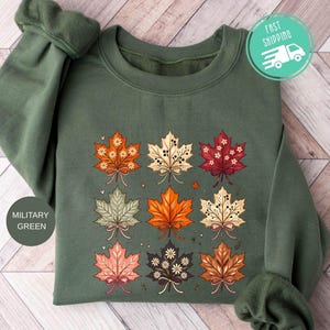 May include: A military green crewneck sweatshirt featuring a graphic print of nine stylized maple leaves arranged in a grid. Each leaf displays unique patterns, including small flowers, dots, and intricate line work, with a small bow at the base.
