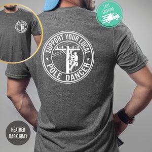 May include: A heather dark gray t-shirt with a white graphic on the back. The graphic is a circle with the text "Support Your Local Pole Dancer" and a silhouette of a person climbing a utility pole.