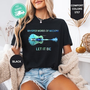 May include: A black short-sleeve t-shirt featuring a graphic of an acoustic guitar formed by trees and a reflection. The text reads WHISPER WORDS OF WISDOM and LET IT BE in blue and white. The shirt includes a Comfort Colors 1717 label.