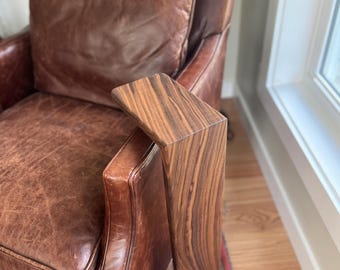 May include: A brown leather armchair with a wooden side table attached to the armrest. The wood has a striped grain pattern. The chair is placed on a patterned rug near a window.