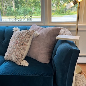 May include: A teal velvet sofa with two decorative pillows. One pillow is a solid light mauve color, and the other is patterned with tassels. A small wooden side table is attached to the sofa's arm.