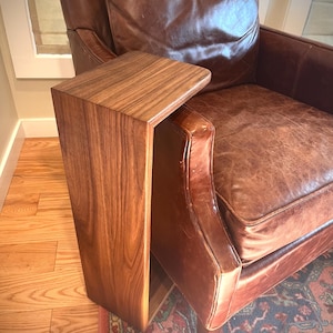 May include: A wooden side table with a warm brown finish. The table is designed to slide over the arm of a brown leather armchair. The wood grain is visible on the table's surface, adding to its natural aesthetic.