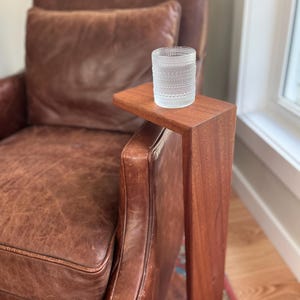 May include: A clear glass tumbler with a textured pattern sits on a small, rectangular wooden side table attached to a brown leather armchair. The table is made of a rich, dark wood. The armchair has a worn, vintage look.