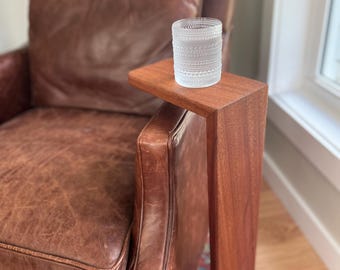 May include: A clear glass tumbler with a textured pattern sits on a small, rectangular wooden side table attached to a brown leather armchair. The table is made of a rich, dark wood. The armchair has a worn, vintage look.