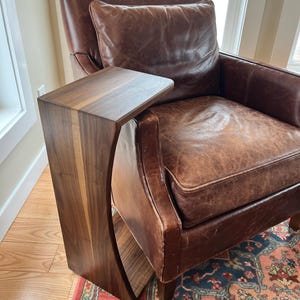 May include: A dark brown wooden side table with a curved design, attached to a brown leather armchair. The table has a rectangular top and a vertical support with a curved cutout. The armchair has a brown leather cushion and pillow.