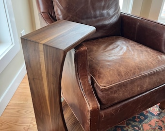 May include: A dark brown wooden side table with a curved design, attached to a brown leather armchair. The table has a rectangular top and a vertical support with a curved cutout. The armchair has a brown leather cushion and pillow.