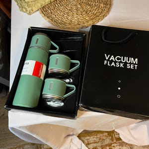May include: A mint green vacuum flask set with two matching mugs and a flask. The set is in a black box with the words "VACUUM FLASK SET" printed on it. The flask has a red and white label that says "Hot & Cold".
