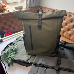 May include: An olive green roll-top backpack featuring a black plastic buckle closure, a vertical zippered front pocket, and side pockets. The backpack is constructed from a smooth, water-resistant material and includes a top carry handle.
