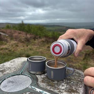 May include: A person pours coffee from a grey and silver flask into a grey mug. Another mug sits nearby. The scene is outdoors with a cloudy sky and a scenic view of the landscape.