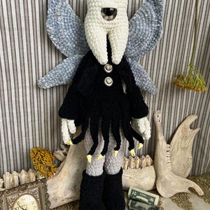 May include: A crochet doll featuring a white tooth-shaped head with a single central eye, large grey textured wings, and a black garment with dangling fringe. The doll has light-colored hands with yellow tips, grey legs, and black boots, standing upright.