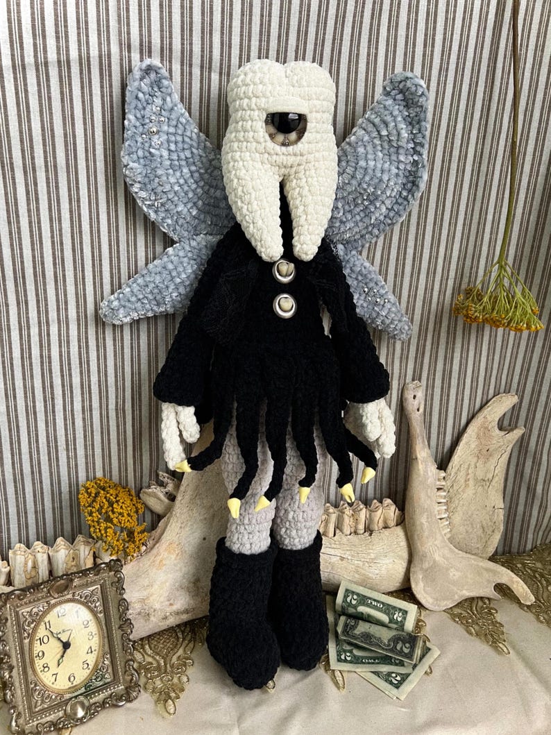 May include: A crochet doll featuring a white tooth-shaped head with a single central eye, large grey textured wings, and a black garment with dangling fringe. The doll has light-colored hands with yellow tips, grey legs, and black boots, standing upright.