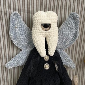 May include: A plush doll featuring a cream-colored tooth-shaped head with a single large central eye, grey velvet-textured wings with silver bead accents, and a black crocheted body adorned with two buttons resembling human teeth.