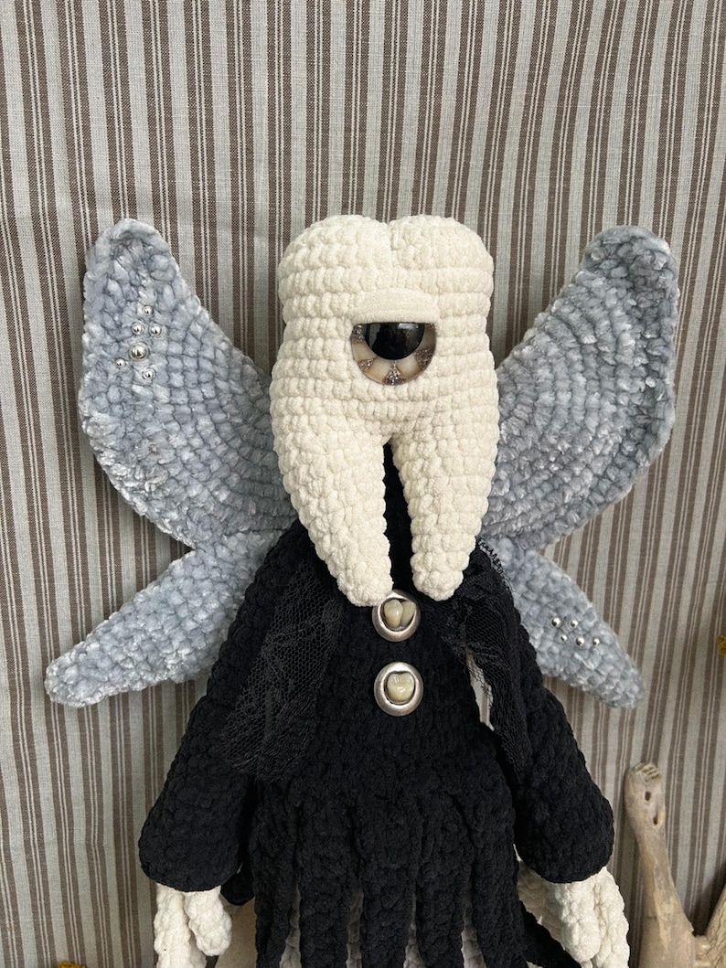 May include: A plush doll featuring a cream-colored tooth-shaped head with a single large central eye, grey velvet-textured wings with silver bead accents, and a black crocheted body adorned with two buttons resembling human teeth.