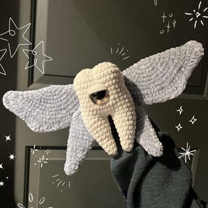 May include: A plush crochet creature featuring a central off-white tooth-shaped body with a single large brown eye and two expansive, textured light blue wings extending from the sides, decorated with white hand-drawn star and sparkle graphics.