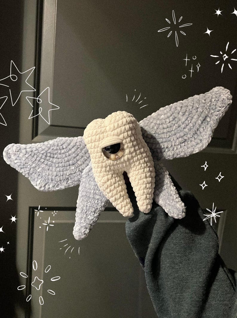 May include: A plush crochet creature featuring a central off-white tooth-shaped body with a single large brown eye and two expansive, textured light blue wings extending from the sides, decorated with white hand-drawn star and sparkle graphics.