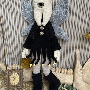 May include: A crochet doll featuring a white tooth-shaped head with a single central eye, large grey textured wings, and a black jacket with two buttons. The doll wears a black fringed skirt, light grey legs, and black boots with yellow-tipped fingers.