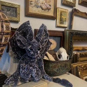 May include: A plush, dark purple, crocheted dragon with large, pointed ears and a long tail. The dragon is sitting on a book with a decorative, ornate, metal bowl behind it.
