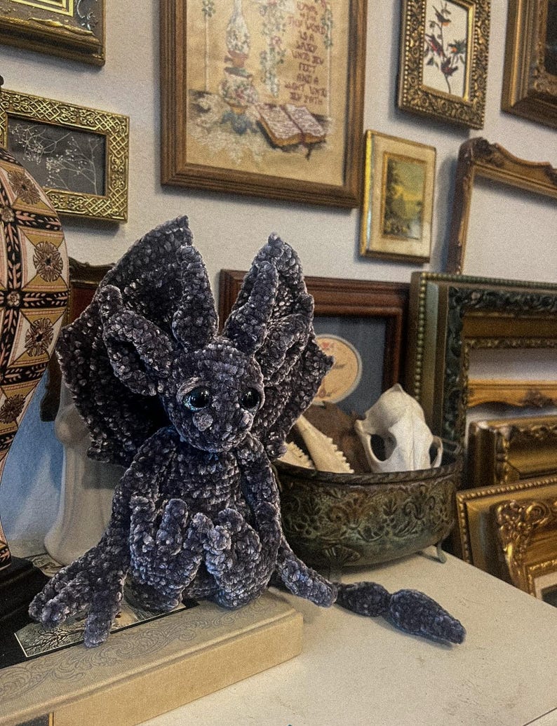 May include: A plush, dark purple, crocheted dragon with large, pointed ears and a long tail. The dragon is sitting on a book with a decorative, ornate, metal bowl behind it.