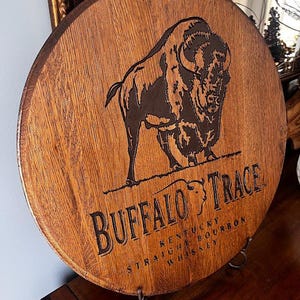May include: Circular wooden sign featuring a dark brown buffalo illustration and the words "BUFFALO TRACE KENTUCKY STRAIGHT BOURBON WHISKEY" in black lettering. The sign is made from a section of a wooden barrel.