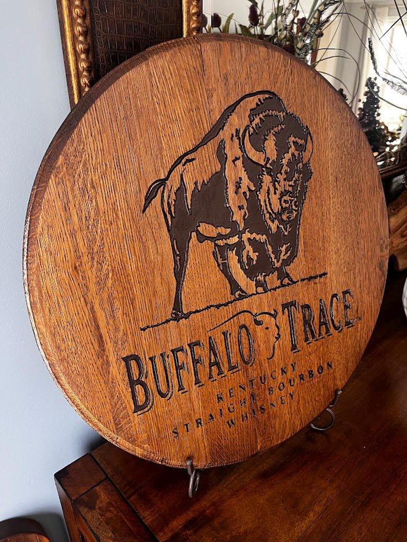 May include: Circular wooden sign featuring a dark brown buffalo illustration and the words "BUFFALO TRACE KENTUCKY STRAIGHT BOURBON WHISKEY" in black lettering. The sign is made from a section of a wooden barrel.