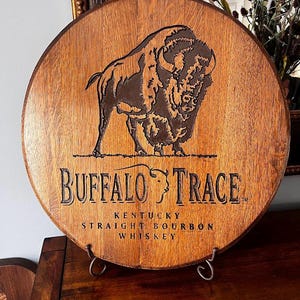 May include: Circular wooden sign featuring the Buffalo Trace logo. The sign displays a detailed engraving of a bison and the words "Buffalo Trace Kentucky Straight Bourbon Whiskey" in black lettering. The sign is displayed on a metal stand.