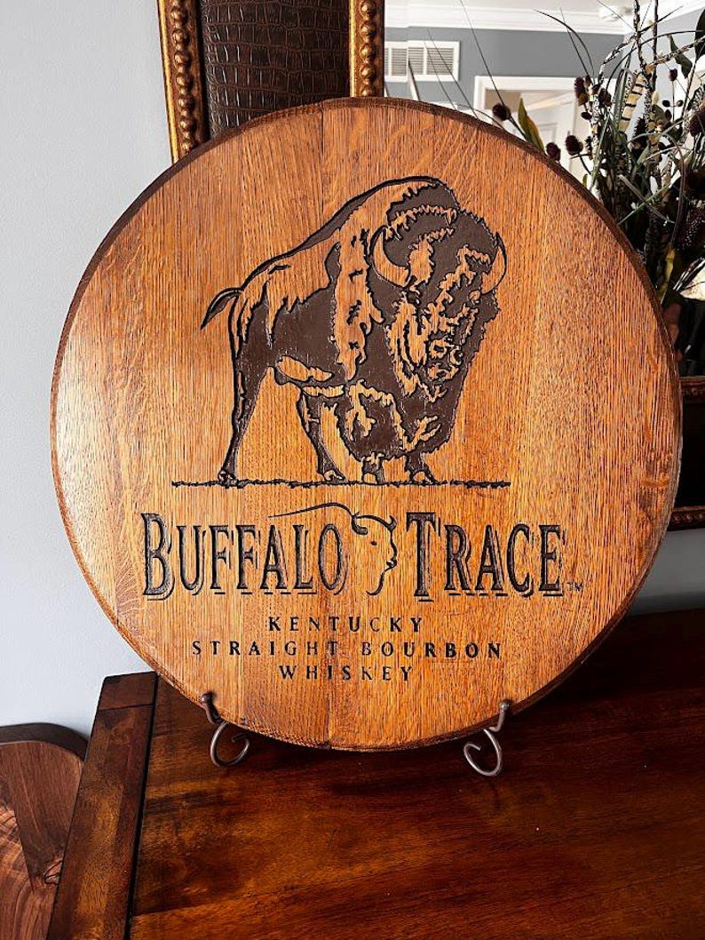 May include: Circular wooden sign featuring the Buffalo Trace logo. The sign displays a detailed engraving of a bison and the words "Buffalo Trace Kentucky Straight Bourbon Whiskey" in black lettering. The sign is displayed on a metal stand.