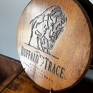 May include: A circular wooden sign featuring the Buffalo Trace logo and text. The sign has a detailed engraving of a bison and the words "Buffalo Trace Kentucky Straight Bourbon Whiskey". It is displayed on a metal stand.