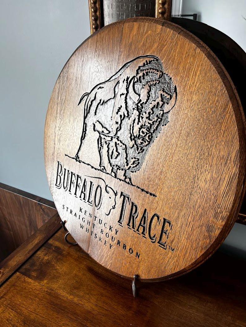 May include: A circular wooden sign featuring the Buffalo Trace logo and text. The sign has a detailed engraving of a bison and the words "Buffalo Trace Kentucky Straight Bourbon Whiskey". It is displayed on a metal stand.