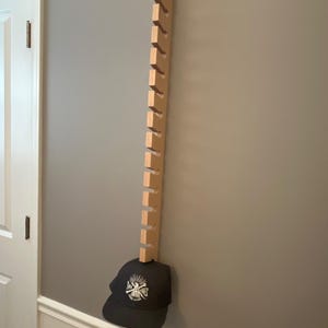 May include: A wooden hat rack with multiple slots is mounted on a grey wall. A dark grey baseball cap with a white graphic is hanging from the rack. The rack is made of light-coloured wood.