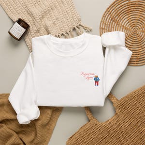 May include: A white long-sleeve sweatshirt featuring a small embroidered graphic on the left chest. The graphic depicts a brown bear wearing a blue jacket and red pants, accompanied by the red cursive text that reads Seguimo Aqui.