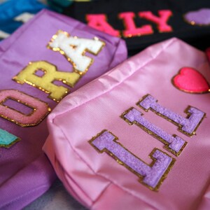 May include: Close-up of several small, colorful bags. Each bag has a name in varsity-style letters. The bags are in shades of pink, purple, blue, and black. Some bags have heart-shaped patches.