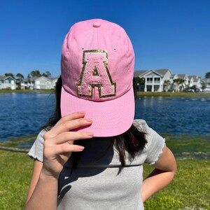 May include: A pink baseball cap with a gold-trimmed, fuzzy letter "A" patch. The cap is made of a soft material and has a curved brim. The hat is a stylish accessory for casual wear.