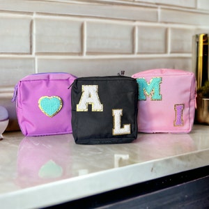 May include: Three small, zippered cosmetic bags in purple, black, and pink. The purple bag has a turquoise heart, the black bag has the letters "AL", and the pink bag has the letters "MI". Each letter is white with a gold border.