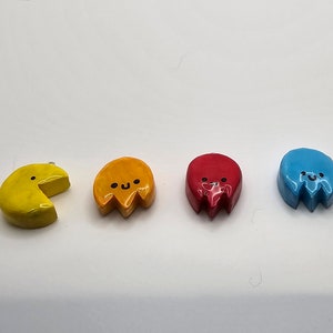 May include: Four small ceramic figurines of Pac-Man characters. The first figurine is yellow and shaped like Pac-Man with a bite taken out of it. The next three figurines are ghosts, one orange, one red, and one blue. Each ghost has a cute face with eyes and a smile.